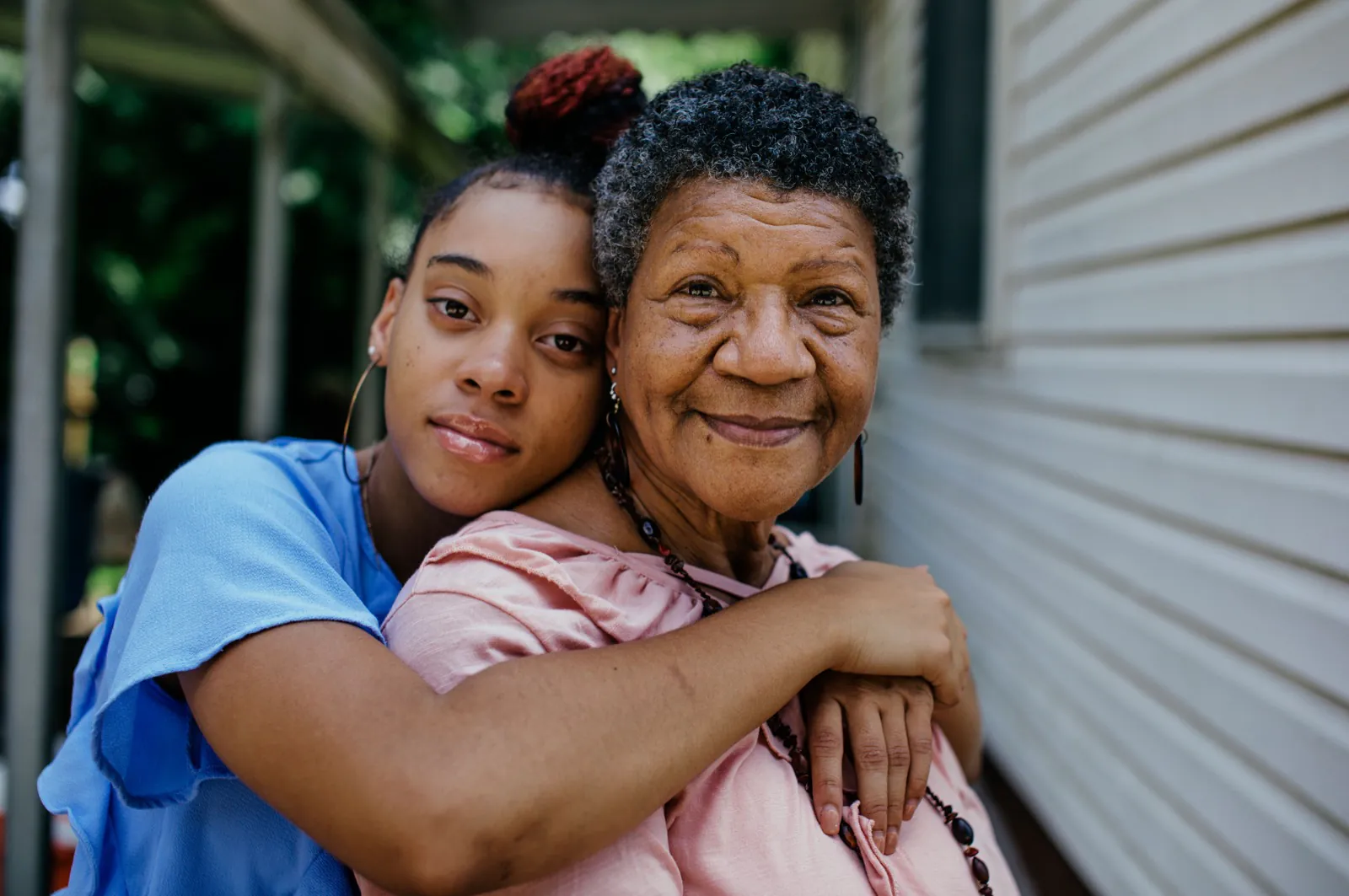 An adult granddaughter wraps her arms around her smiling grandmother on a sunlit Houston front porch.