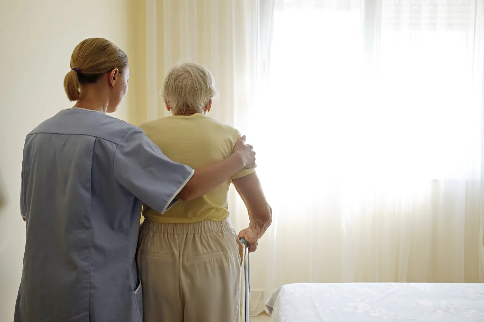 A caregiver steadies an elderly person with a cane as they walk toward a sunlit window.