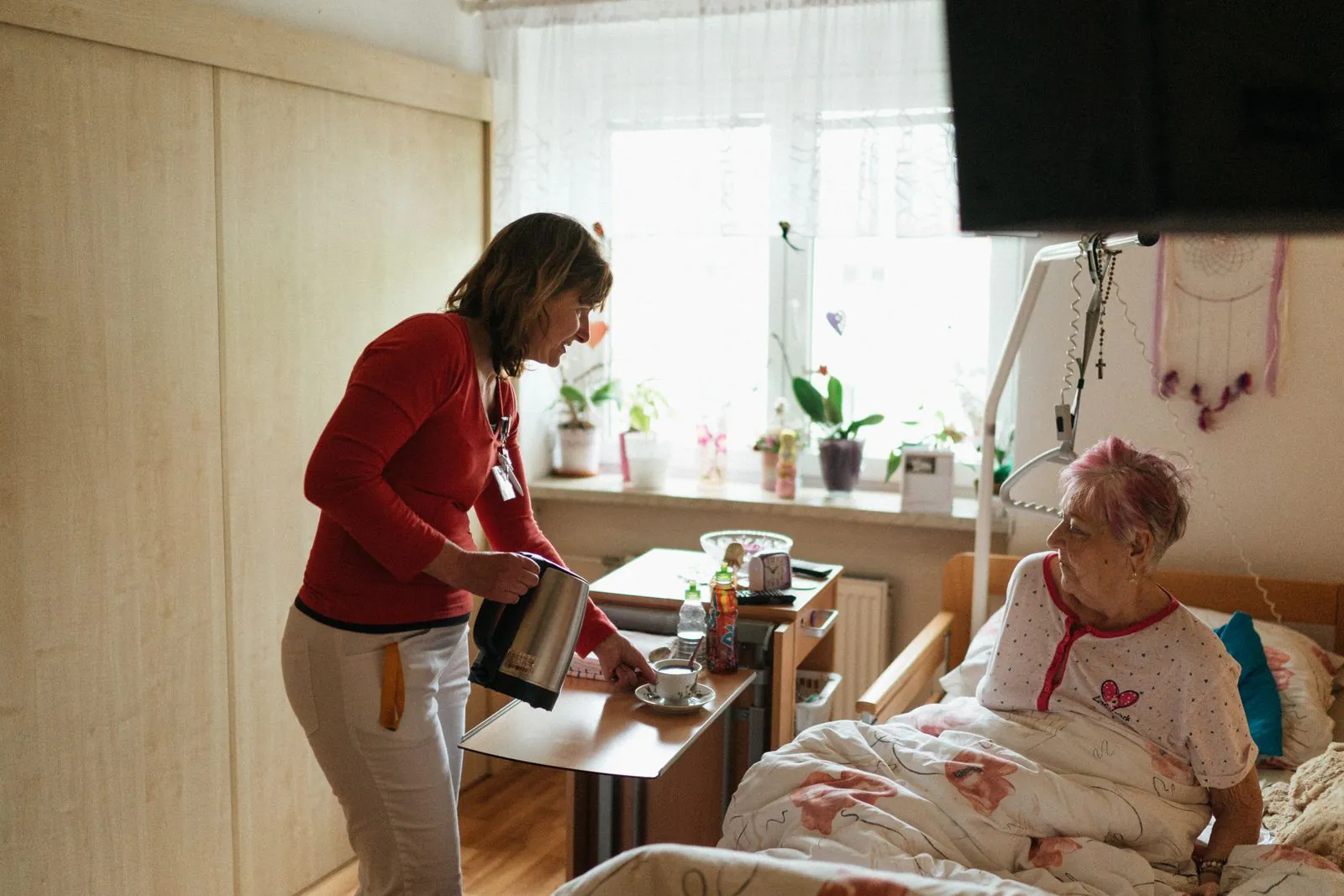 A caregiver pours tea at the bedside of an elderly woman in a sunlit bedroom.